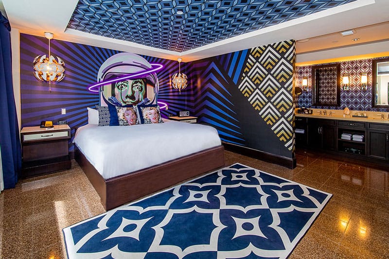 A wide-angle view of a hotel room with a large bed, nightstands, a vanity area, and a decorative ceiling.