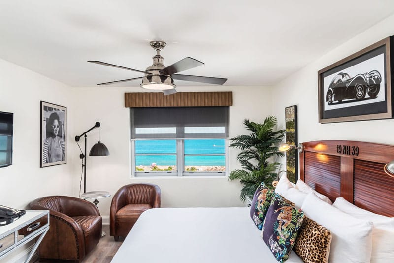 A hotel room with a large bed, two brown leather armchairs, a ceiling fan, and a window with a view of the ocean.