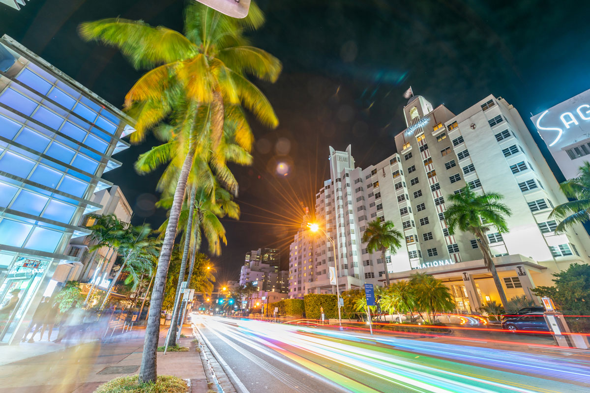 A nighttime view of a hotel exterior with palm trees and city street