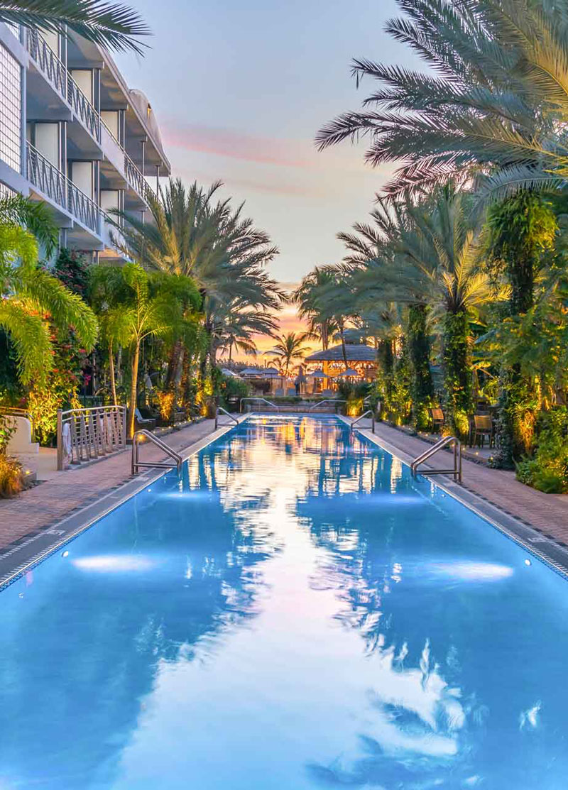 a long rectangular pool surrounded by palm trees and a multi-story building with balconies at sunset