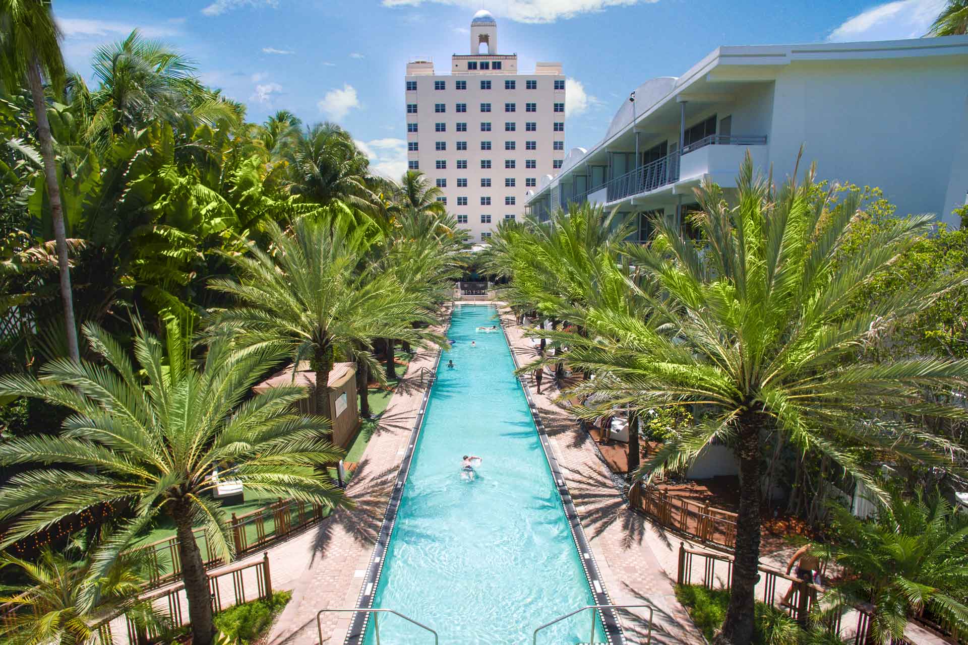 A long pool surrounded by palm trees with a hotel building in the background.
