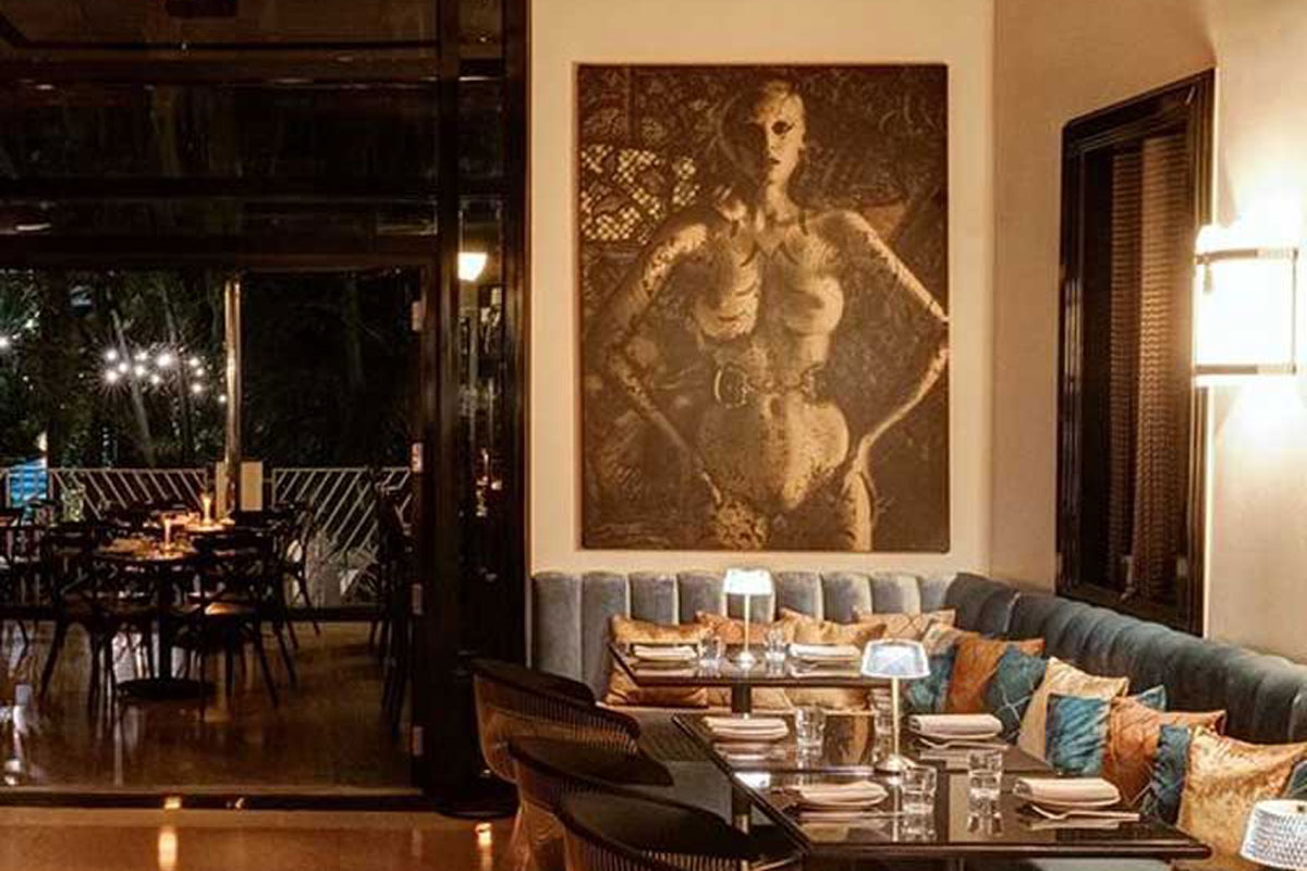 A dining area with a booth, tables, and chairs, featuring a large artwork on the wall and glass doors leading to an outdoor seating area.
