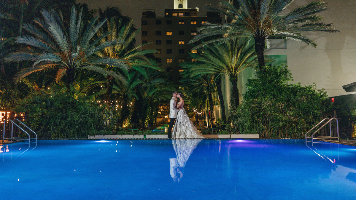 A couple in wedding attire embracing by the pool at nighttime, surrounded by palm trees and string lights.